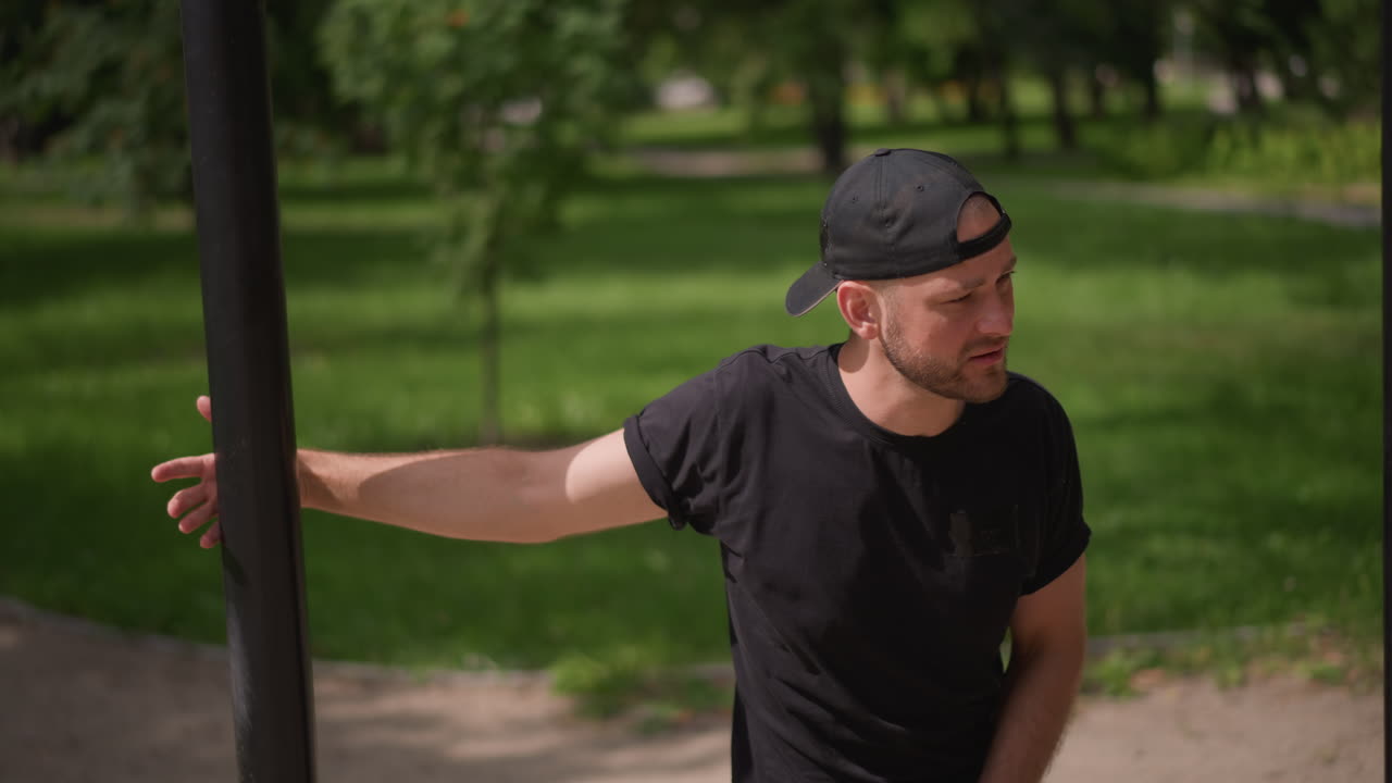 man stretching arm on pole, outdoor warmup and recovery scene showing man leaning against street pole, extending arm across body with slight squint from sun, casual cap and tshirt, leafy path