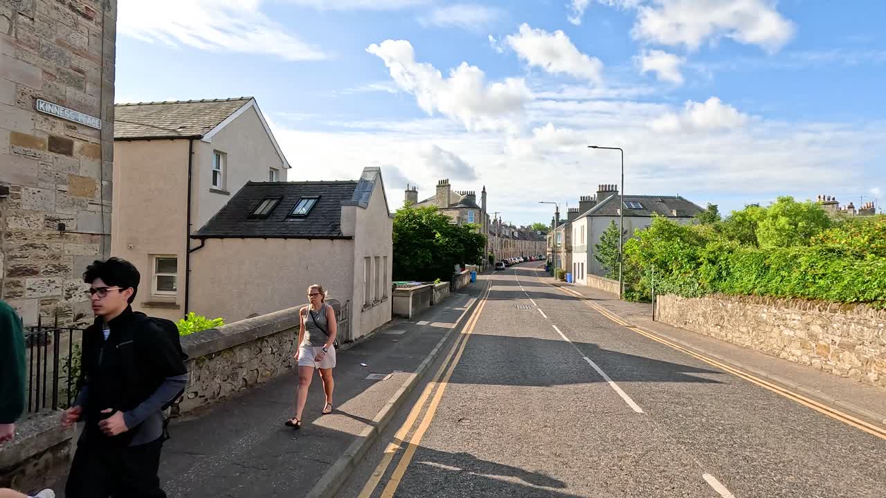 Daytime street view of stone buildings, pedestrians, parked cars, and blue sky in Fife
