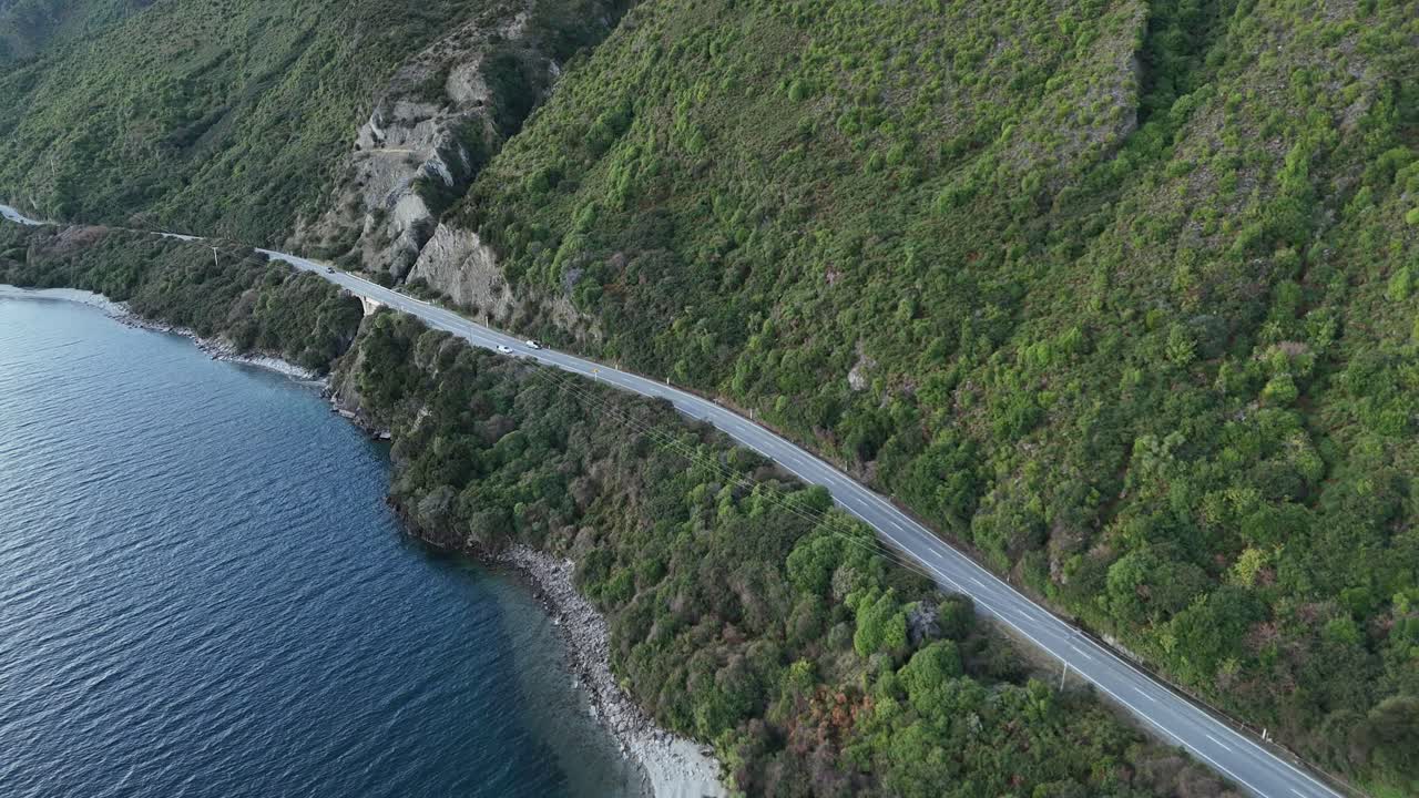 Drone flying over a scenic highway running between a crystal-clear lake and towering green mountains
