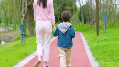 Loving-Mother-Meets-Her-Son-From-Classroom,-Holds-For-Hand-And-Taking-To-Home-Passing-Through-Green-Park-In-Morning-Sunny-Day