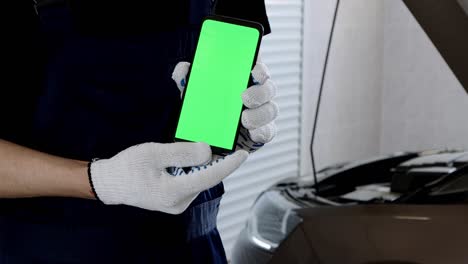 un trabajador de garaje sostiene un teléfono con una pantalla verde en el fondo de un capó abierto del coche. reparación de equipos y motor en un taller de automóviles.