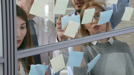 equipo de negocios de mentores senior y colegas jóvenes escribiendo y pegando notas adhesivas en la pared de vidrio en la oficina y hablando de planes