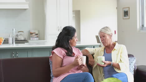 Felices-Y-Diversas-Mujeres-Mayores-Con-Café,-Riéndose-En-Un-Sofá-En-Una-Soleada-Sala-De-Estar,-Cámara-Lenta