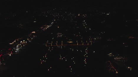 Dark-night-river-city-aerial:-Coloured-lights-move-far-below,-pattern