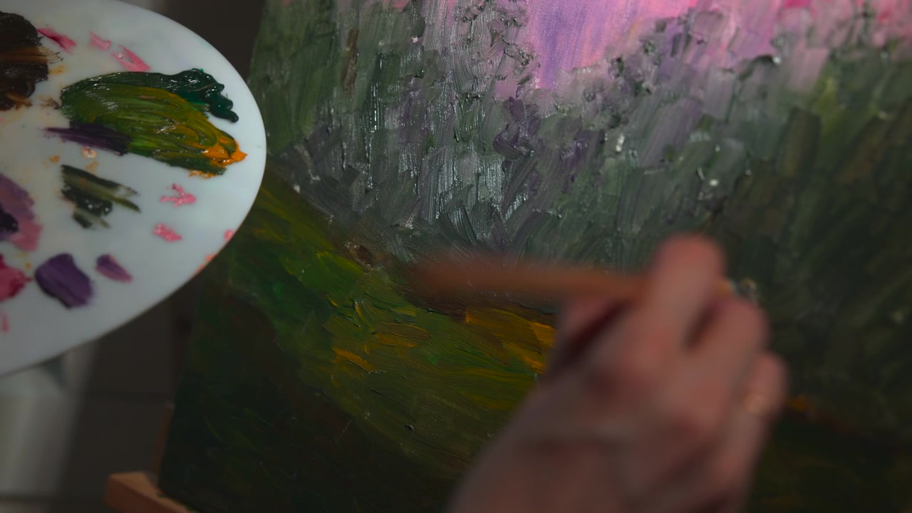 Close-up of a hand using a brush to paint a textured abstract canvas beside a palette, with soft pink and green tones visible under dim studio lighting