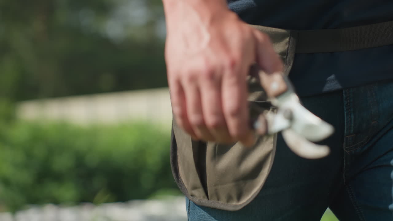 Close up of farmer walking outdoors holding metal plier in hand, tool belt strapped around waist, blue jeans and green garden background visible