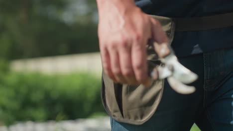 close up of farmer walking outdoors holding metal plier in hand, tool belt strapped around waist, blue jeans and green garden background visible