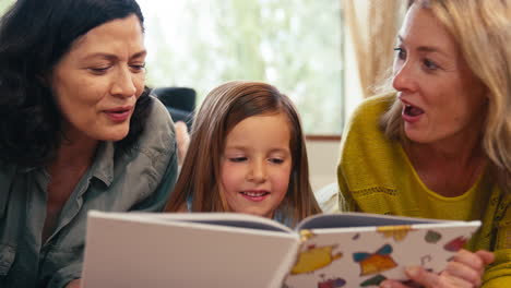 familia del mismo sexo con dos madres e hija acostadas en el suelo leyendo un libro juntos en casa