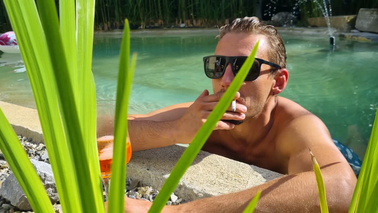 A stylish man in sunglasses relaxes in a turquoise pool in Switzerland, sipping a cocktail and smoking a cigarette, surrounded by lush tropical plants, radiating summer vibes on a hot day