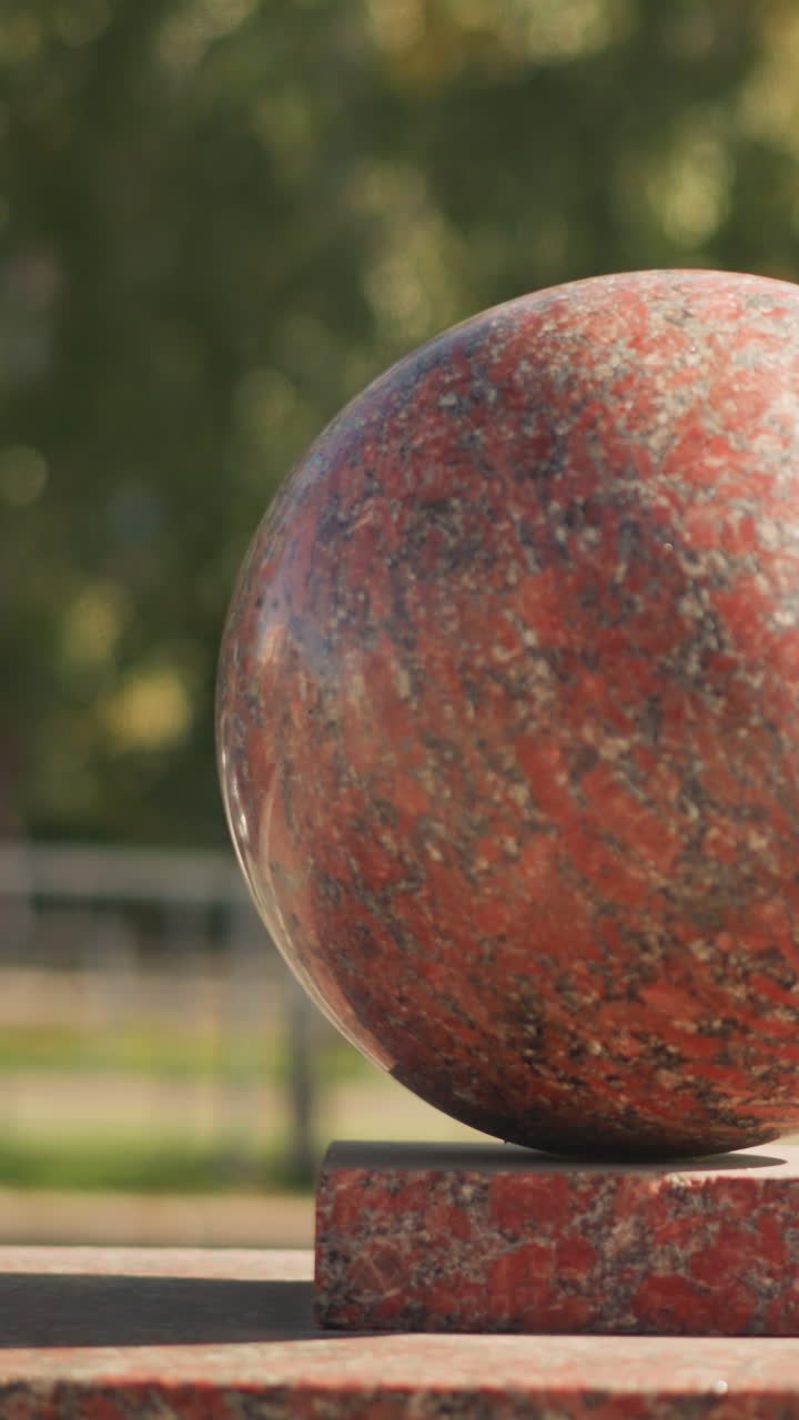 esfera de granito rojo en un camino de parque, piedra moteada sobre un pedestal cuadrado, árboles frondosos y bokeh de césped en el fondo, luz solar suave que proyecta sutiles reflejos sobre la superficie texturizada, verano tranquilo