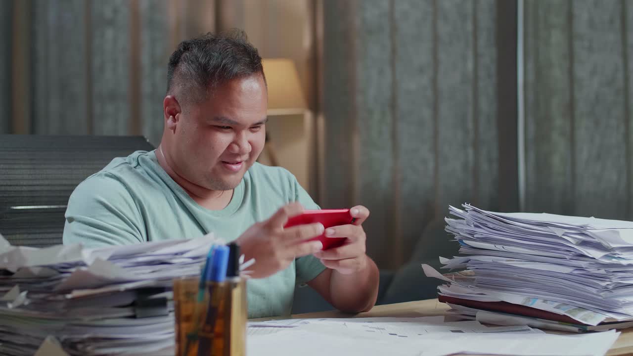 Nærbillede af glad kraftig asiatisk mand, der fejrer at have vundet et spil på smartphone efter at have arbejdet med dokumenter på kontoret.