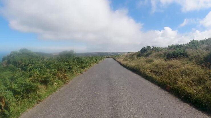 POV di guida in auto su strada di campagna con mare, brughiera verde e cielo azzurro nuvoloso, North Devon, Regno Unito, 4K.