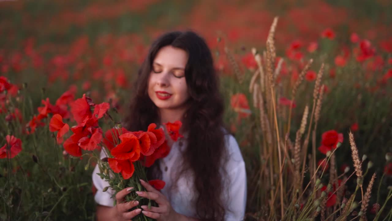 hermosa chica de cabello oscuro en un campo de flores silvestres y amapolas rojas, con un sombrero y un vestido, sosteniendo un ramo de flores y sonriendo cerca de tallos de trigo