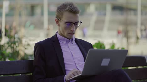 Concentrated-young-man-sitting-on-bench-and-typing-on-laptop