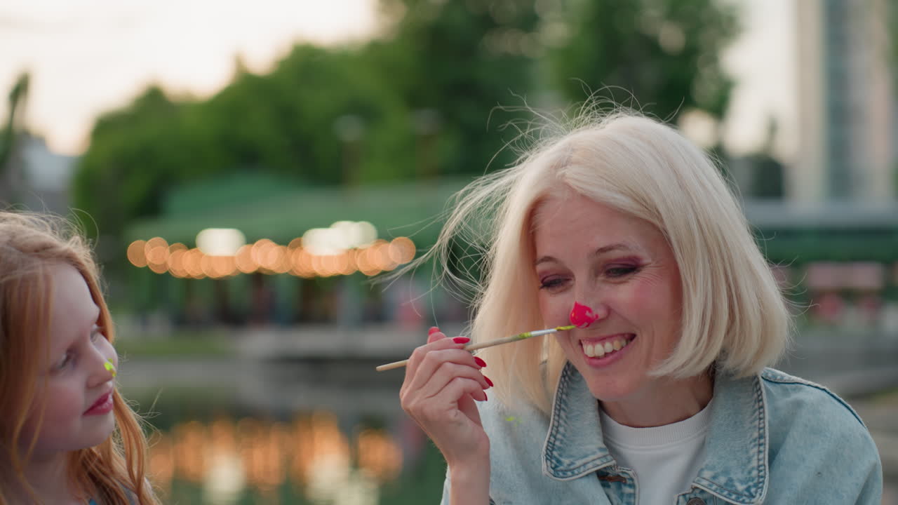 joyful mom with bright paint on nose giggles as young daughter playfully presses paint brush against mom nose during warm sunset painting session by lakeside park, sharing creative family moment