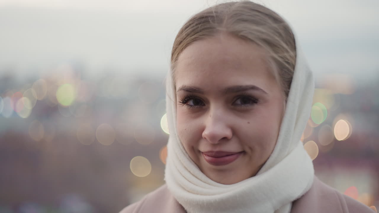 Primo piano nitido di una giovane donna con la sciarpa su un tetto, con bokeh morbido. Lo sguardo attento si trasforma in un sorriso caloroso, con luci serali sfocate dietro di lei. Una pendolare, avvolta nel cappotto e nella sciarpa, fa una pausa speranzosa tra gli impegni.