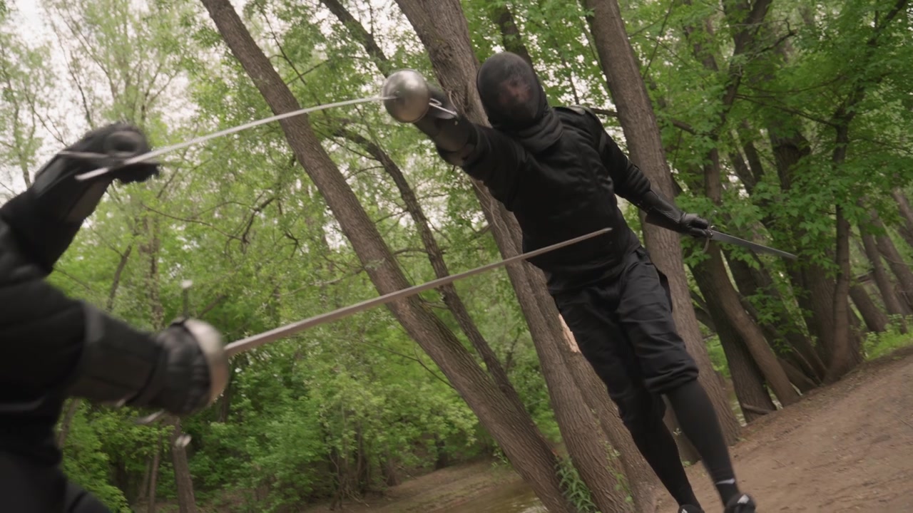 Two men in black martial arts gear engage in sword fight along forest path demonstrating precise movements and techniques. scene features dense green foliage surrounding action with natural