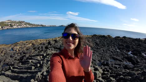 Girl-taking-a-selfie-waving-at-the-camera-near-the-sea-in-slow-motion