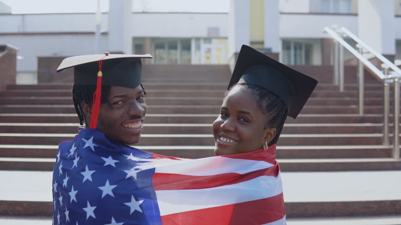 hombre y mujer afroamericanos de pie uno al lado del otro con las espaldas hacia la cámara en sombreros cuadrados de estudiantes graduados con la bandera de los estados unidos en los hombros. mirando a la cámara