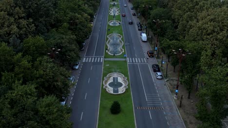 Langsame-Luftaufnahme-Des-Parlamentspalastes-In-Bukarest,-Rumänien-Mit-Einem-Orangefarbenen-Himmel-Im-Hintergrund-Bei-Sonnenuntergang,-üppiger-Vegetation-Und-Wasserfontänen-Darunter,-Langsame-Rückwärtsbewegung
