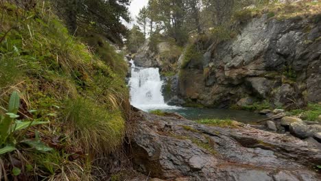 Descansando-Y-Contemplando-La-Orilla-De-Un-Río-De-Montaña-Con-Una-Hermosa-Cascada-Al-Fondo,-Durante-Una-Caminata-Explorando-Y-Descubriendo-Lugares-Escondidos-Y-Remotos