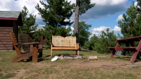 low campfire burning during the day - surrounded by empty muskoka chairs, benches, and log cabins