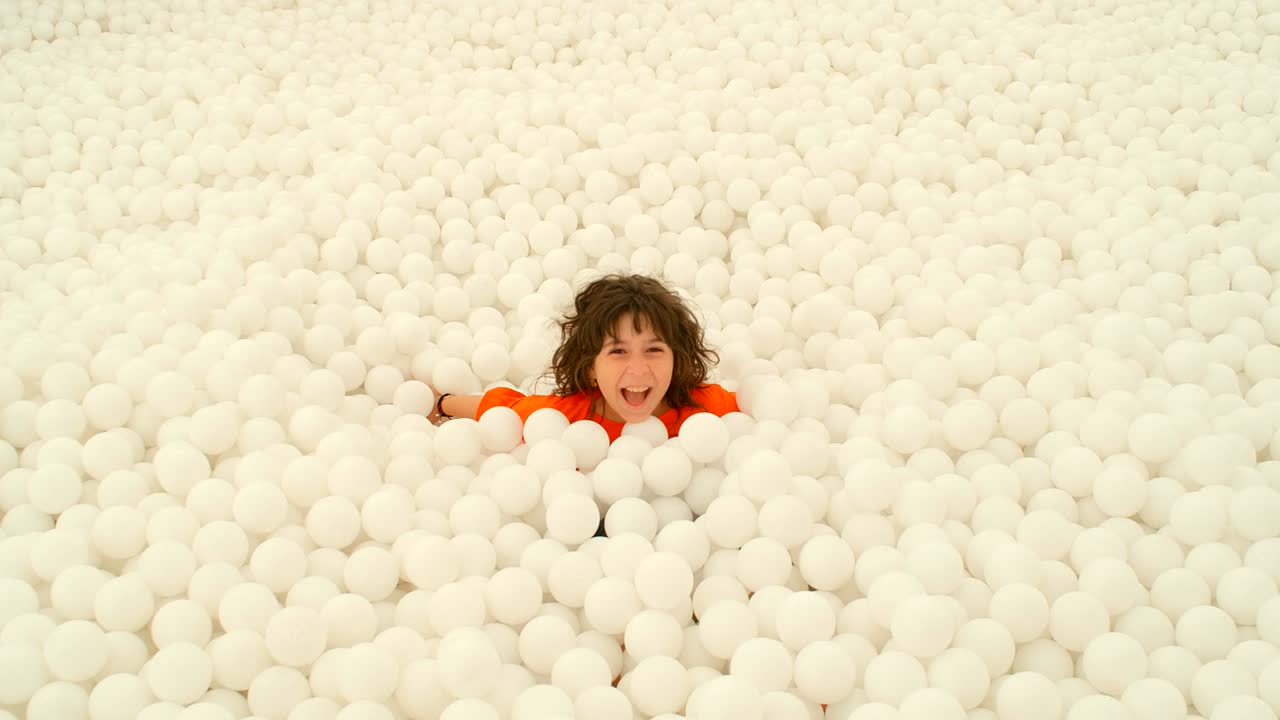 una linda niña de 9-10 años divirtiéndose entre un montón de bolas blancas en un pozo de bolas. 4k cámara lenta