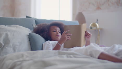 niñita negra con pijama blanco acostada en la cama y leyendo un libro