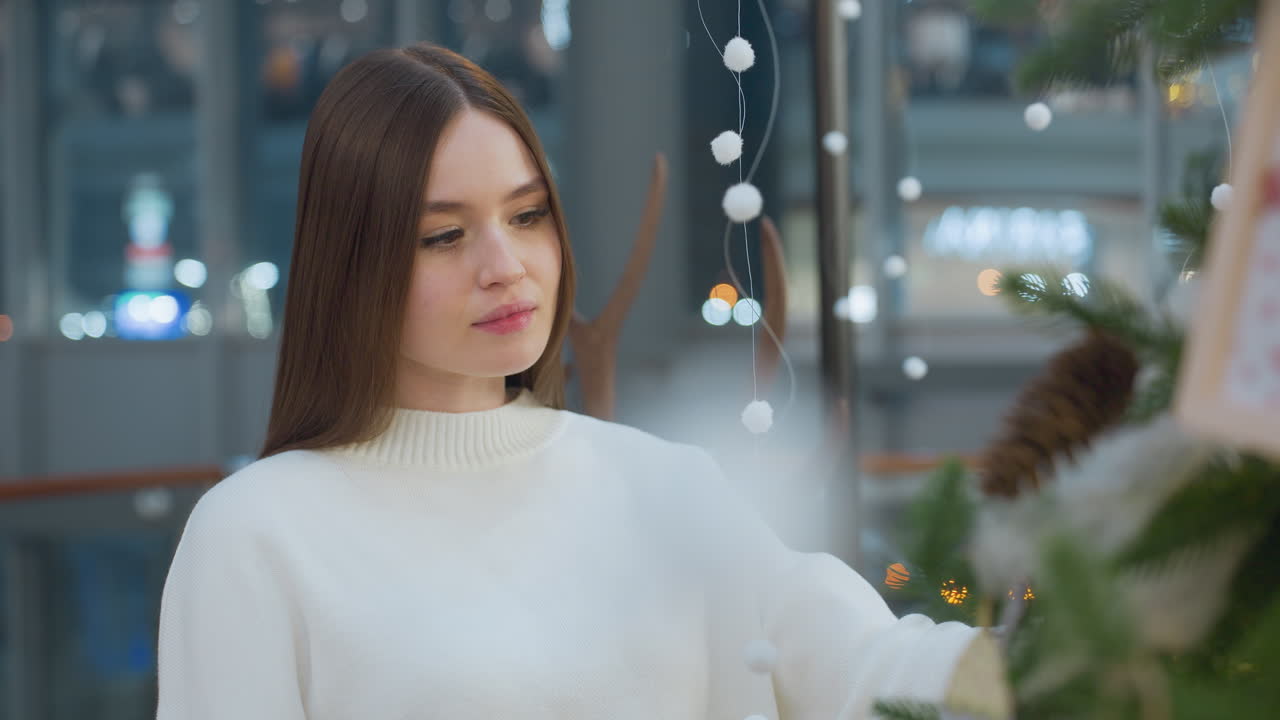 Serene young woman in white sweater admires festive decorations at a shopping mall, exuding a calm holiday spirit with bokeh light in the surrounding