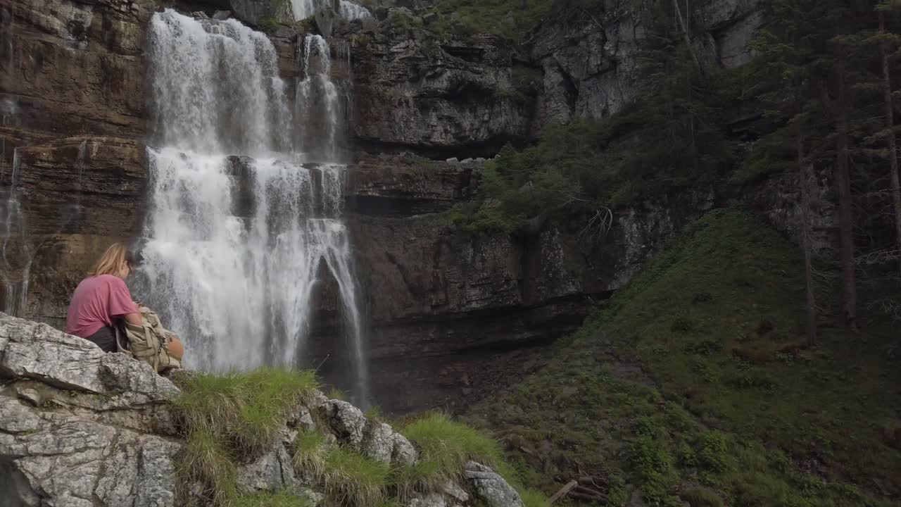 una chica de excursión está sentada en una roca frente a una cascada en la reserva natural