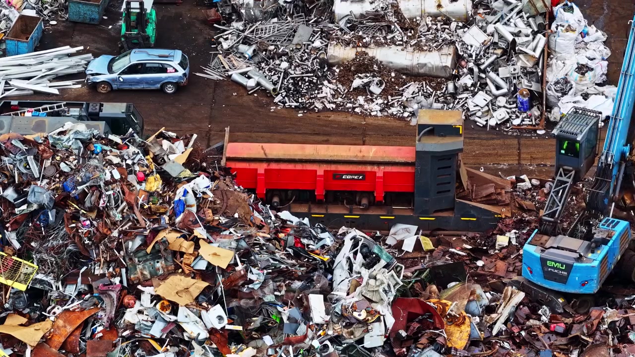 Escavatore carica rottami metallici in un container rosso in un trafficato cantiere di riciclaggio industriale