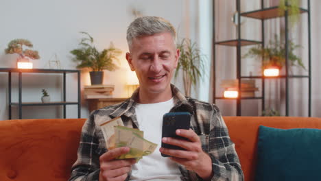 smiling happy caucasian man counting money euro cash use smartphone income salary lottery game win