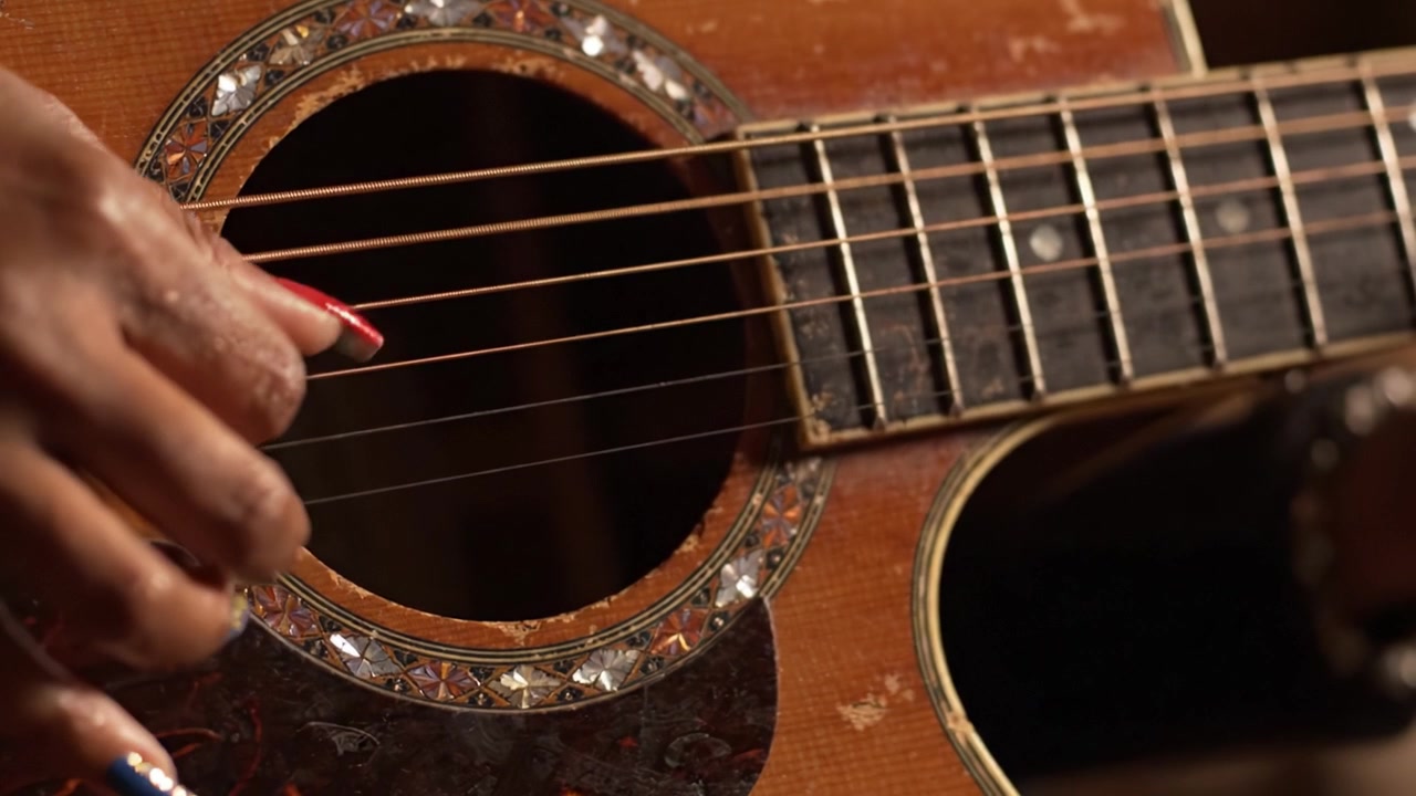 Un primo piano affascinante di una mano che strimpella una chitarra acustica, mostrando i dettagli intricati e le ricche texture dello strumento in un ambiente caldo e accogliente