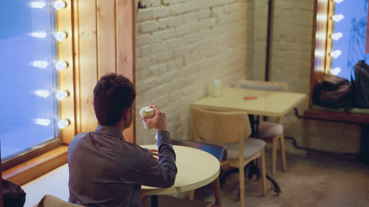 Student wearing formal shirt drinks coffee while sitting alone in cozy cafe, surrounded by wooden chairs, jacket draped over seat, warm lights glowing near window, with peaceful atmosphere and urban decor