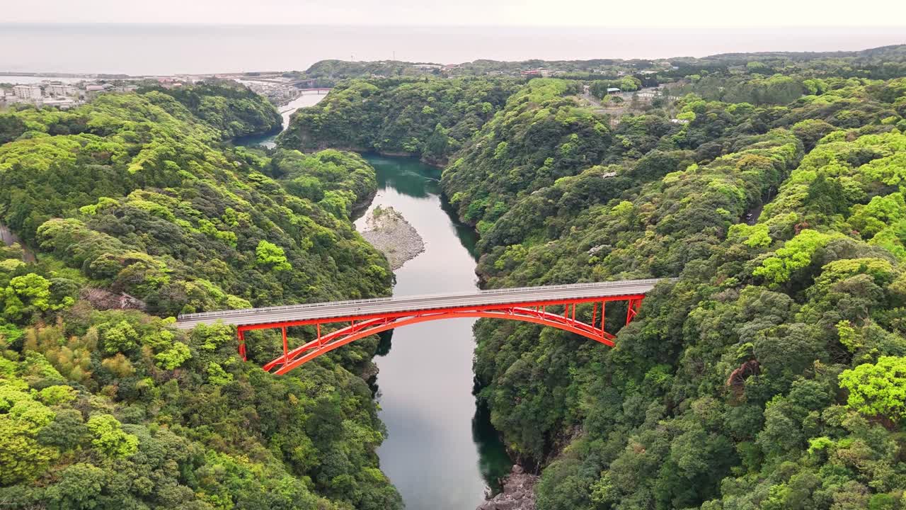 Matsumine Bridge spanning river gorge with red structure contrasting verdant forest landscape in Yakushima, UNESCO World Heritage Site capturing natural infrastructure integration, drone push in