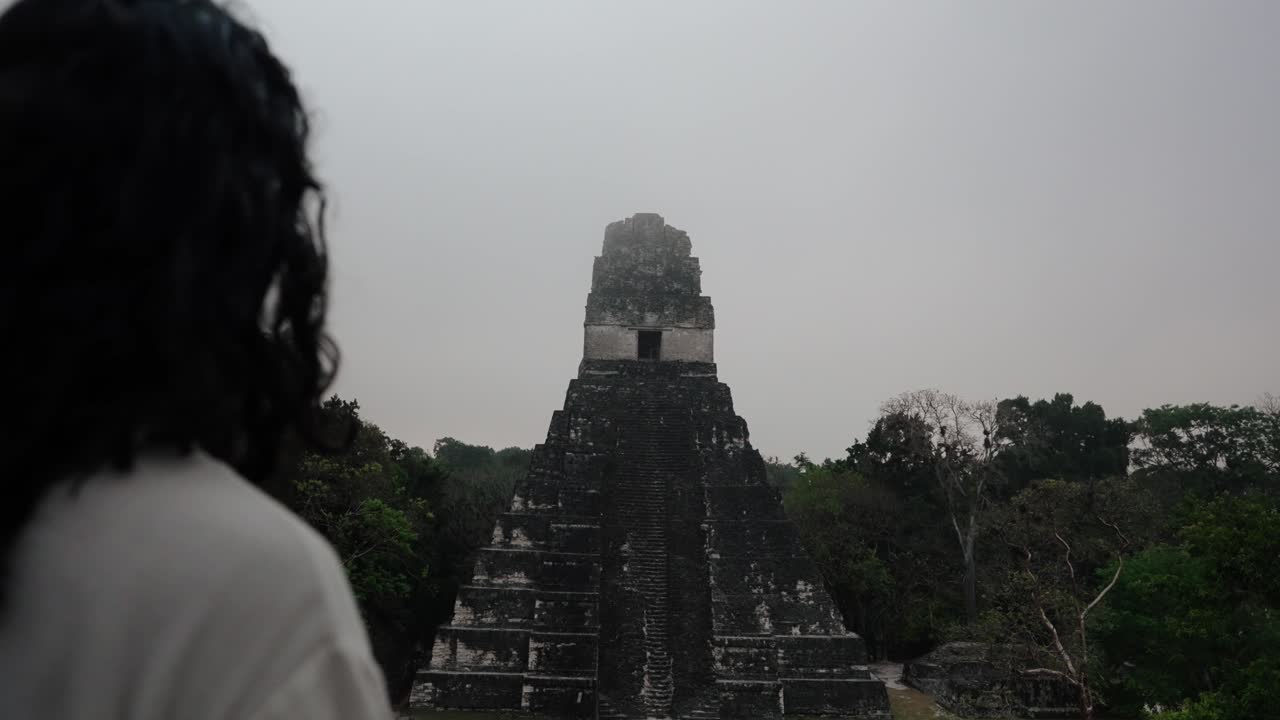A man with long hair stands facing the ancient Mayan pyramid Temple I at Tikal, surrounded by dense jungle.