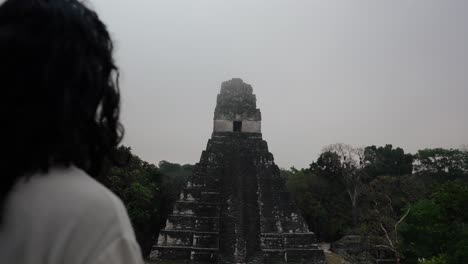 a man with long hair stands facing the ancient mayan pyramid temple i at tikal, surrounded by dense jungle.