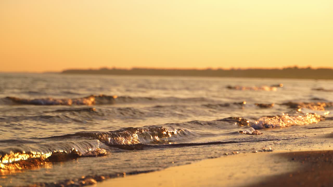 Sea water splashes on sand beach in the evening. The edge of the ocean with calm waves on the shoreline at sunset. Close-up.