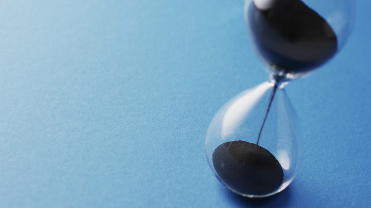 Video of close up of hourglass with black sand pouring with copy space on blue background
