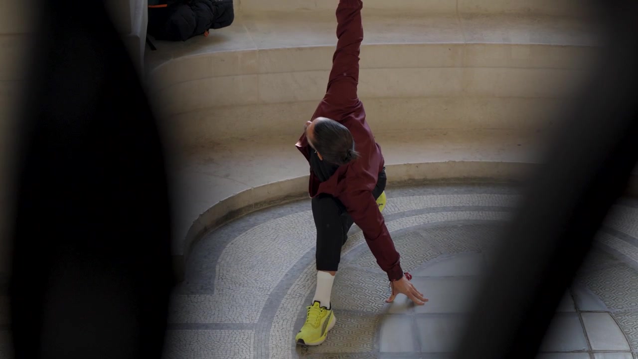 Scatto creativo dall'alto attraverso una struttura in primo piano che mostra un atleta maschio con una giacca bordeaux che esegue affondi dinamici con rotazione in una sala di pietra.