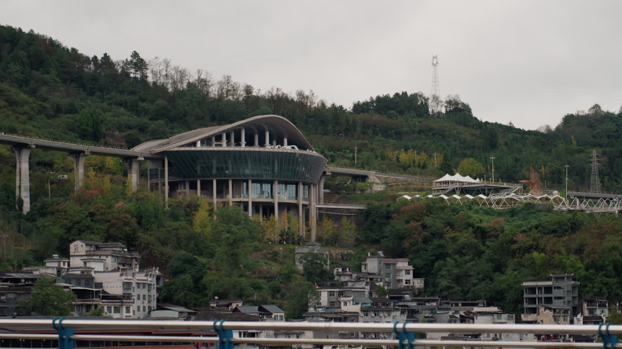 Autobus che passa su un ponte contro una collina, un autobus pubblico bianco attraversa l'inquadratura con un moderno terminal in cemento e case a schiera su un pendio verde, cielo nuvoloso, atmosfera di transito urbano che evoca azioni immaginate.