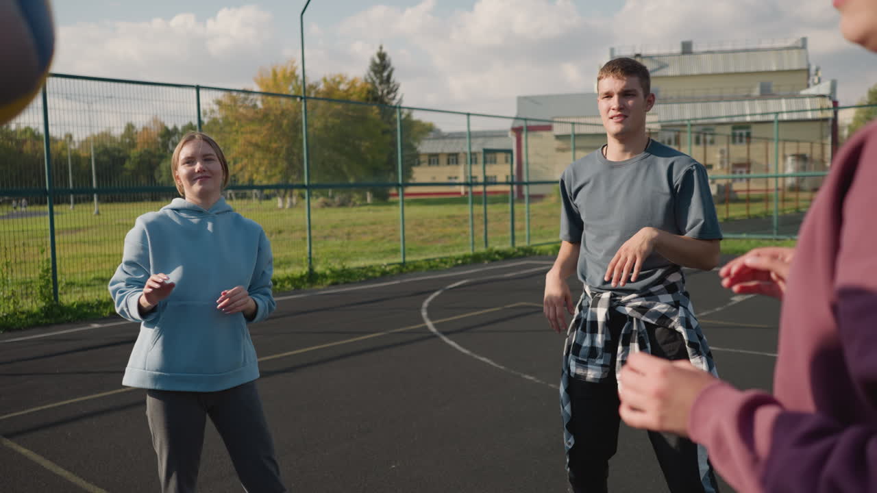 Close-up partial view of person in maroon hoodie passing volleyball to teammate in cyan hoodie while man watches them and reaches out to touch ball during outdoor training session