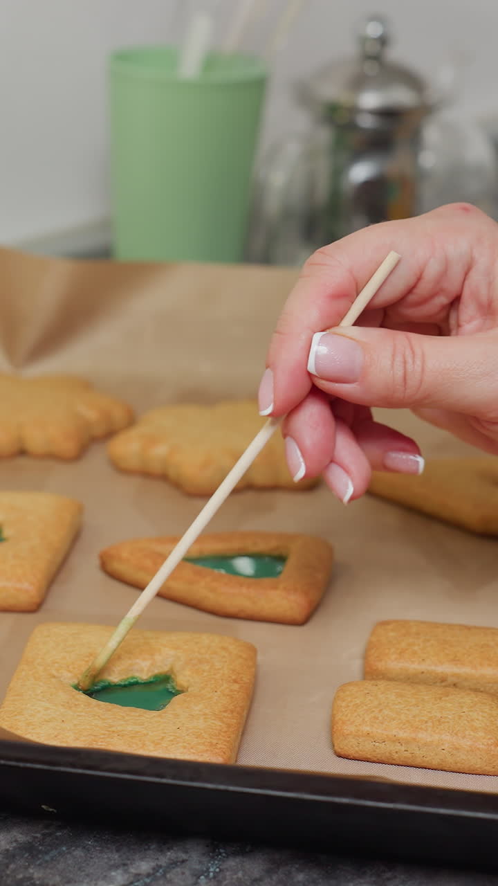 Caterer uses a stick to carefully adjust green liquid in cookie shapes, ensuring precise decoration, other baking equipment, including jars of colorful beads, are visible around the cookie