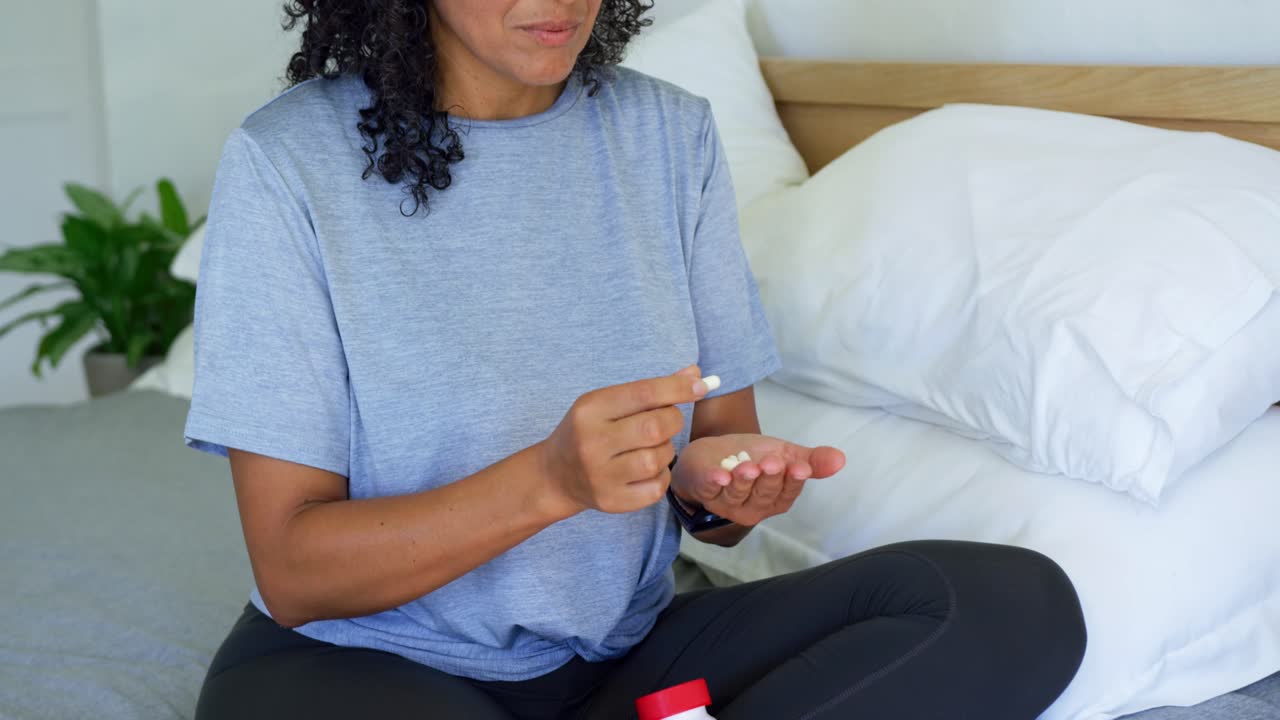 Donna afroamericana che tiene una capsula, seduta sul letto, apre un flacone di pillole e prepara i farmaci.