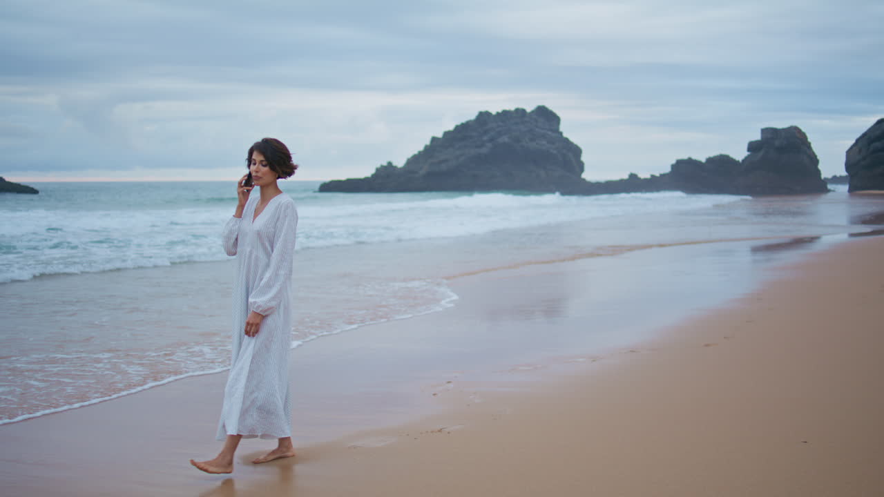아름다운 여자가 바다 해변에서 스마트폰으로 전화합니다. 편안한 여자가 모래 해변을 고 있습니다.