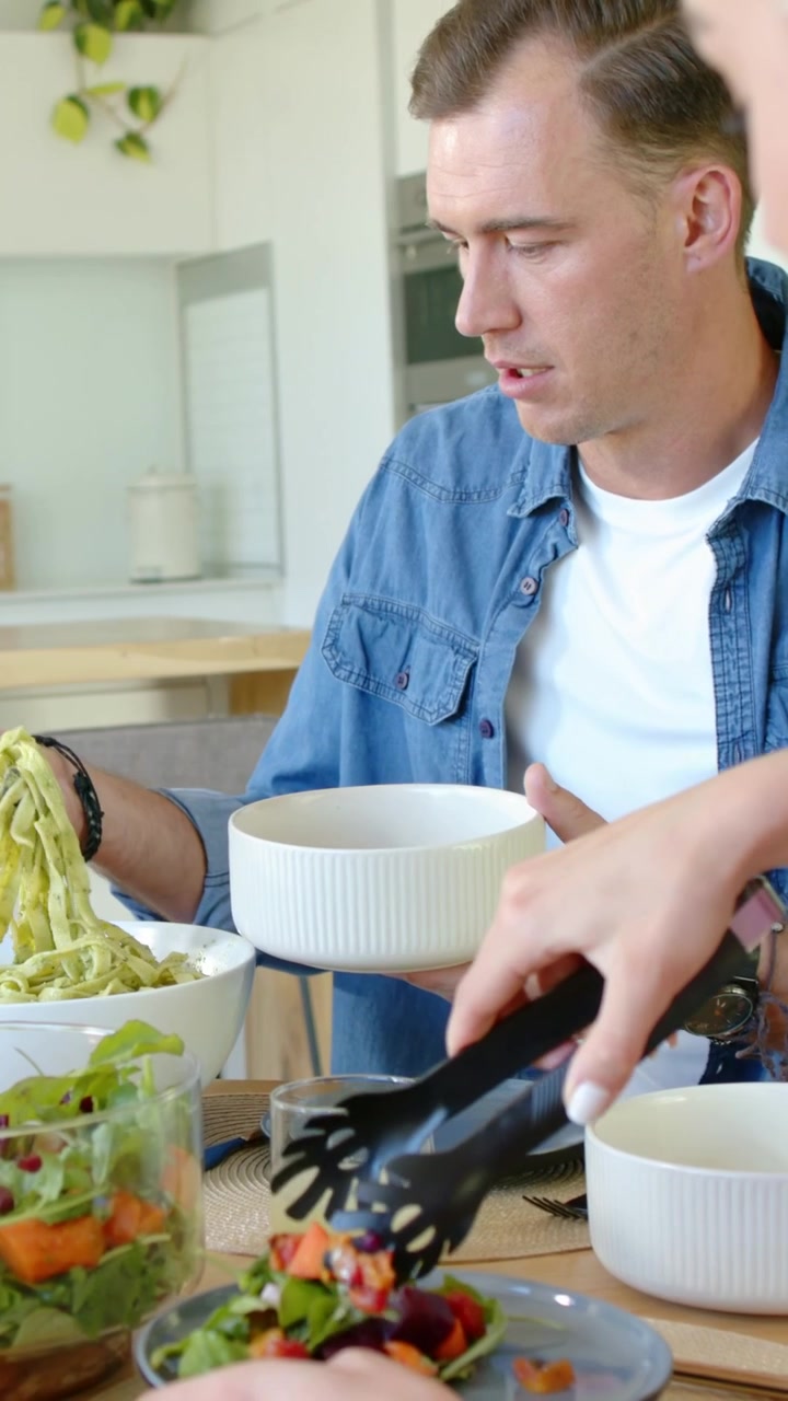 Vertikal video: Holder hvid skål derhjemme, mand i denim tager salat/pasta, serverer med tang
