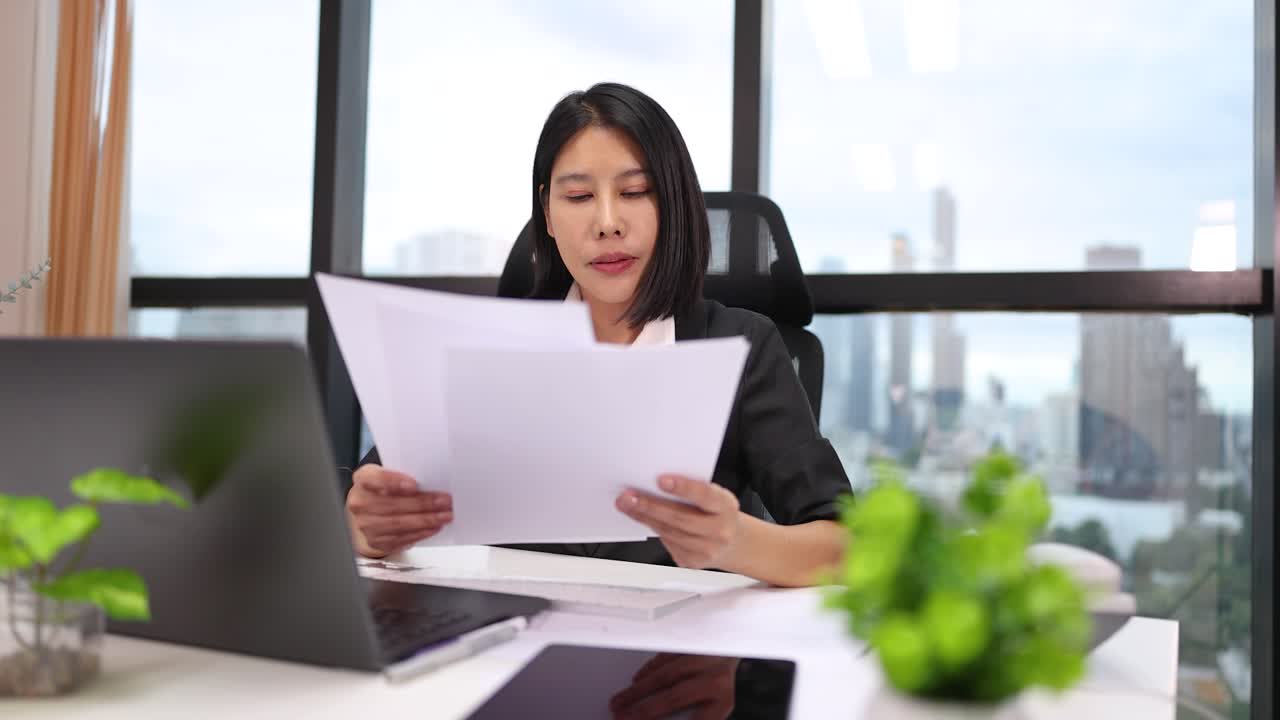 Professional woman examines paperwork at bright office desk with laptop, natural light, city view
