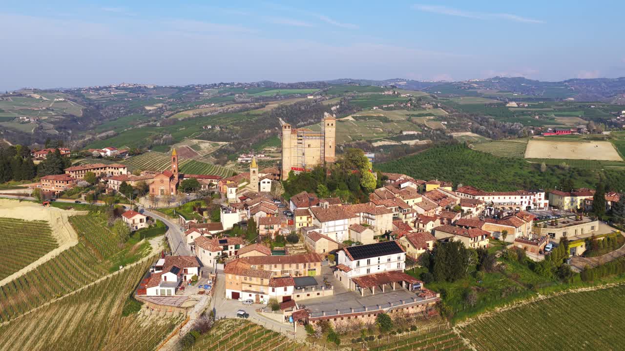 Orbiting aerial shot of Serralunga d'Alba, featuring its iconic medieval castle, charming village rooftops, and lush vineyards of the Langhe region under a clear blue sky