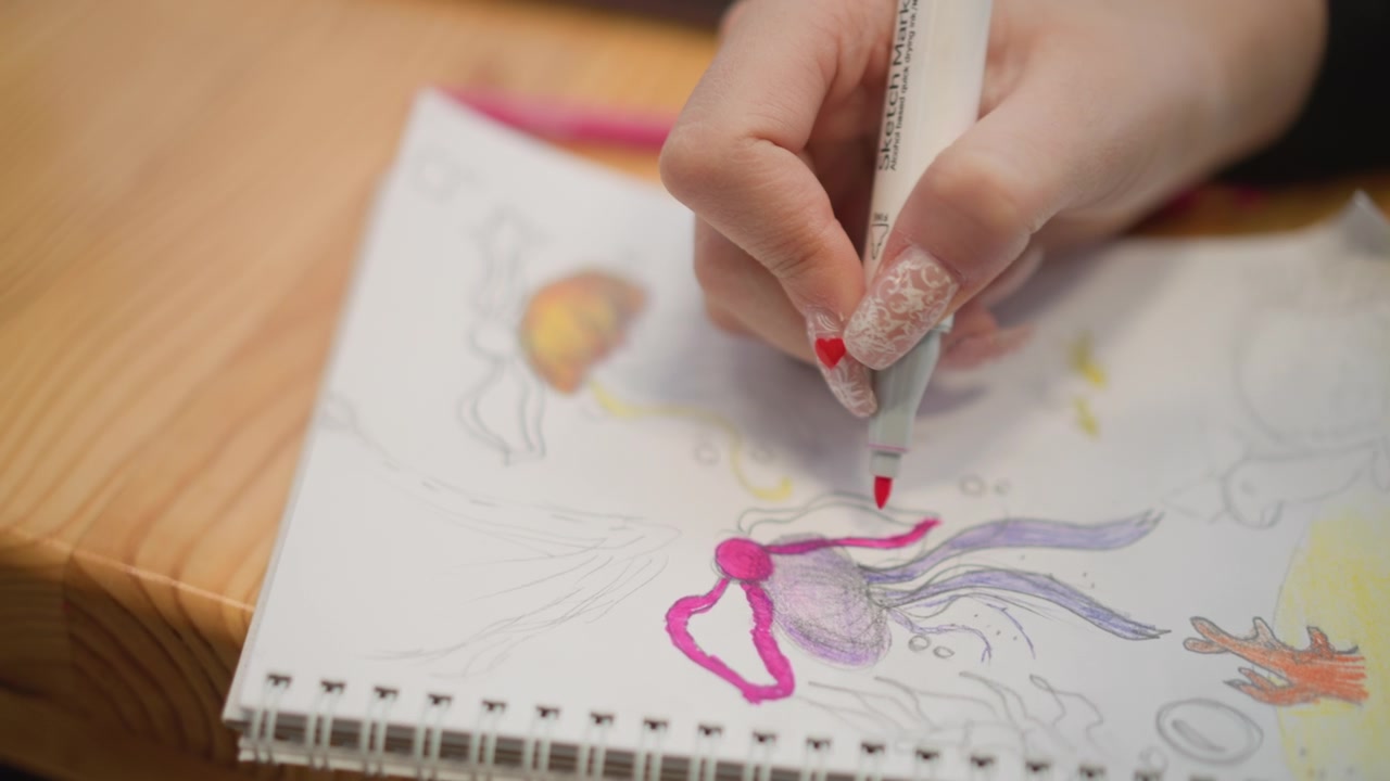 Mano che usa gomma e matita per rimuovere e ridisegnare segni di matita su una pagina di un quaderno a spirale riempita di scarabocchi colorati, inclusi cuore, medusa, fiamma e forme scarabocchiate, sotto una luce calda