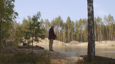 Ein-Einsamer-Mann-Fischt-An-Einem-Sonnigen-Morgen-Am-Ufer-Eines-Flusses-Im-Wald-Und-Ruht-Sich-Im-Urlaub-Aus,-Während-Er-Mit-Der-Rute-In-Der-Hand-Steht